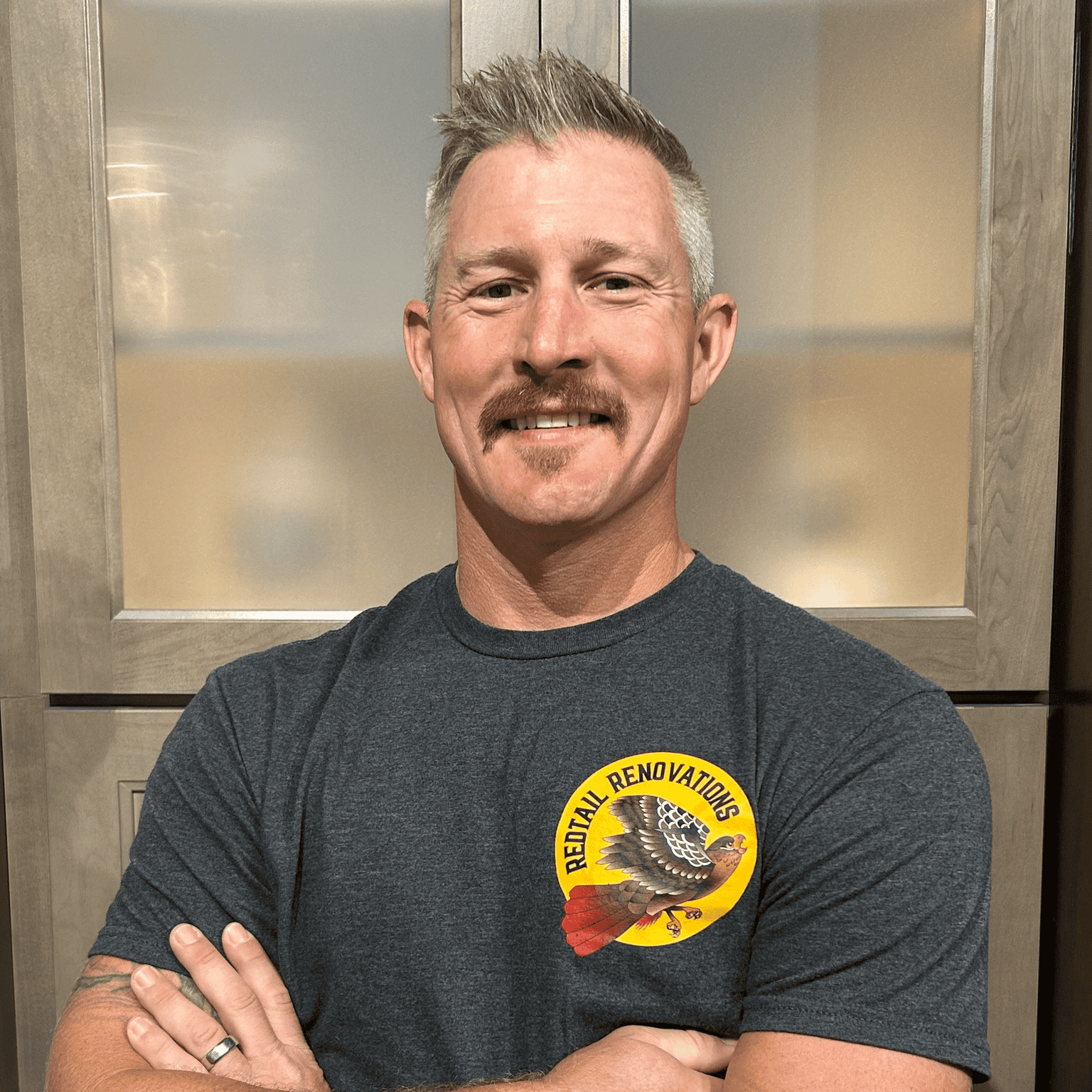 Example Co Team Member Smiling man in a gray T-shirt with 'Rental Renovations' logo, arms crossed, in front of a wooden and.